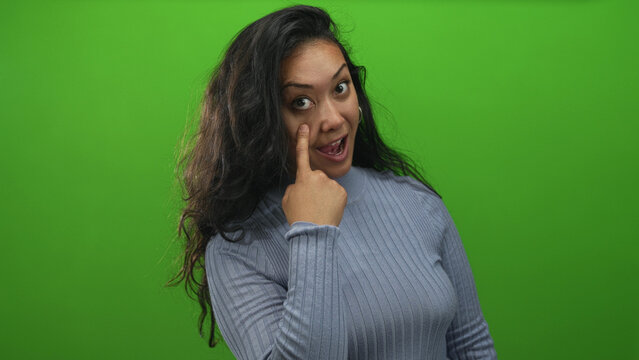 Woman points finger to eye in studio green screen, smirk with raised eyebrow and casual sweater; playful curiosity.