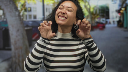 Woman with hands curled in claw gesture grinning on a busy city street in striped top; playful joy...