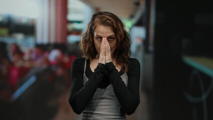 Woman praying intently on restaurant terrace with blurred urban background showcasing outdoor cafe setting under city skyline.