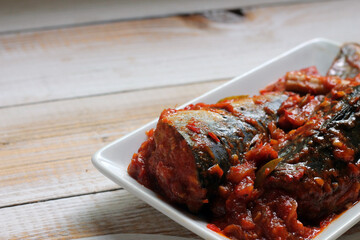 Sarden or Sardencis. Spicy Indonesian mackerel dish served in rich red chili sauce with lemongrass on a white plate over rustic wooden table.