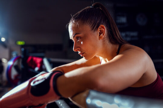 Female boxer leaning against the ropes of a boxing ring resting after training.