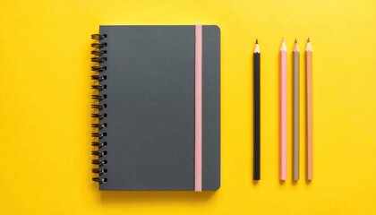 Flat lay of a closed dark grey spiral notebook with a pink elastic band next to 4 pencils on a bright yellow background