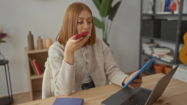 Young blonde woman seated at wooden table holding smartphone to mouth recording voice message in living room; communication concentration connection.