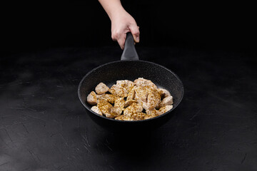 Hand holding a frying pan filled with seasoned chicken pieces, showcasing the cooking process against a dark textured background, emphasizing culinary skills and preparation