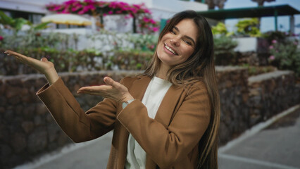 Young hispanic woman smiling and pointing with hands on a vibrant city street surrounded by colorful flowers and outdoor settings wearing a stylish coat in the afternoon light.