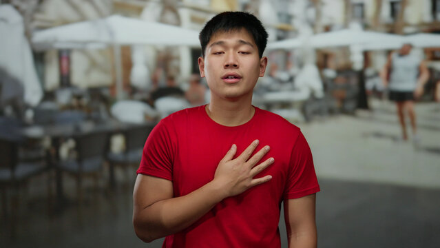 Young chinese man in red shirt gestures emotionally on outdoor terrace of a bustling coffee shop with blurred people in background, capturing urban social ambiance