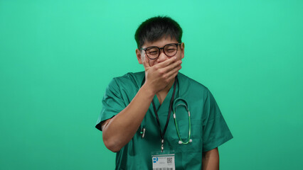 Young chinese man in a green medical uniform laughing joyfully over an isolated green background...