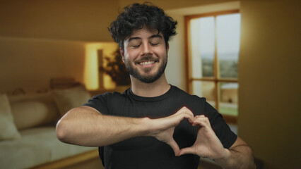 Young man making heart symbol with hands in cozy hotel room with large window and warm lighting,...