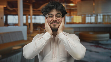 Obraz premium Young hispanic man with beard and glasses stands indoors at a hotel, smiling with hands on face, wearing a white shirt, in a relaxed and joyful ambiance.
