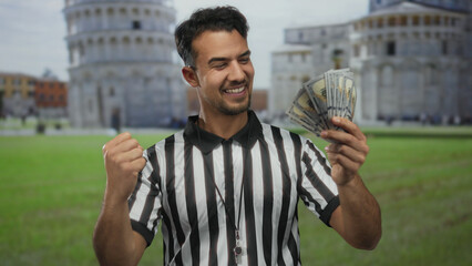 Young hispanic man in referee uniform holding us dollars with pisa tower in the background, smiling...