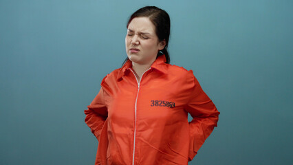 Woman wearing orange prison jumpsuit standing against blue background with a pained expression and...