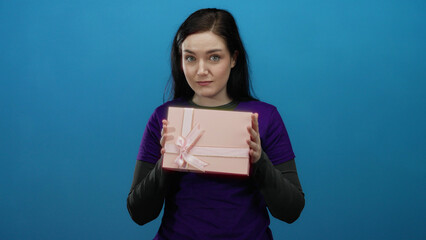 Obraz premium Woman holding pink gift box in front of blue background wearing purple volunteer shirt with joyful expression.