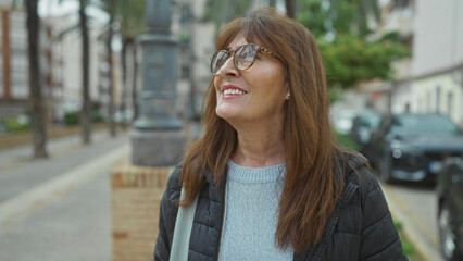 Fototapeta premium Senior hispanic woman with glasses smiling outdoors on a city street showing contentment and serenity in her expression as she enjoys urban life and surrounding environment