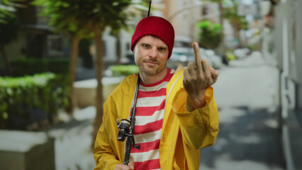 Young man displaying middle finger in urban street with fishing rod and yellow jacket, red beanie, and striped shirt. © Krakenimages.com