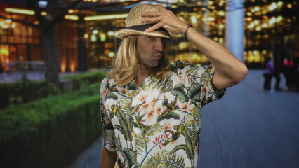 Man tilting straw hat with hand over his face on city street sidewalk against illuminated building...