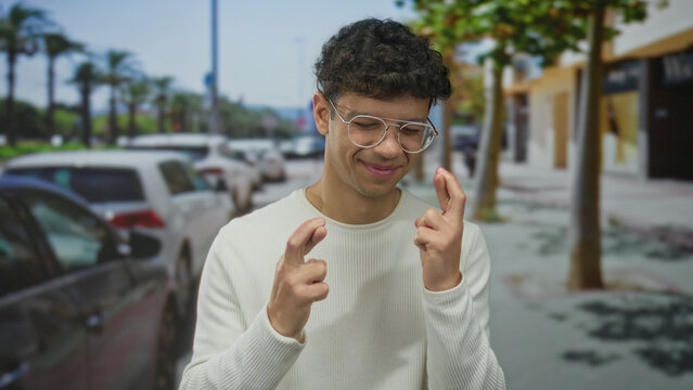 Young hispanic man wearing white sweater and glasses lightly crosses fingers with both hands while standing on a sunlit urban street; hope. - Powered by Adobe