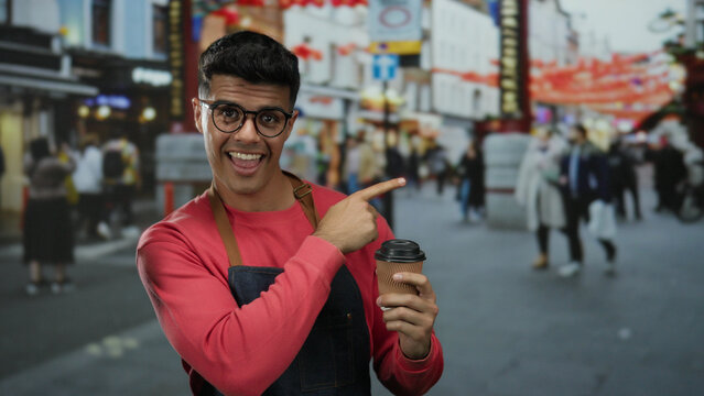 Young man with glasses holding coffee cup on bustling city street surrounded by vibrant urban setting and diverse crowd.