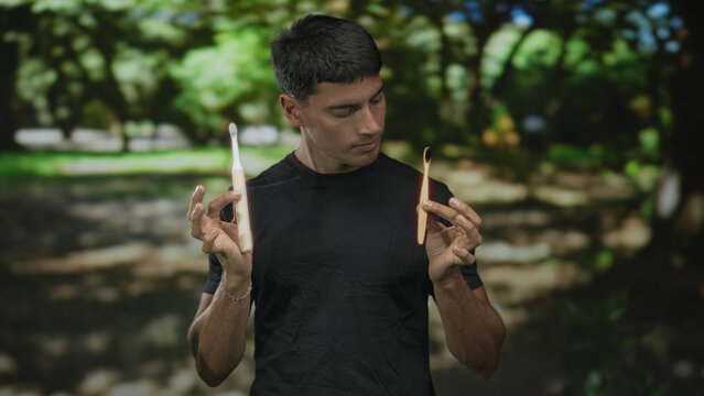 Young hispanic man holds electric toothbrush and bamboo toothbrush side by side in green park; sustainable choice.
