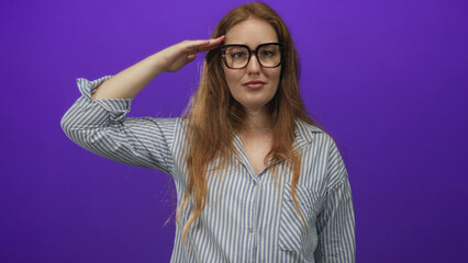 Woman with long red hair in striped shirt and glasses salutes with right hand against purple studio...