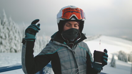 Young woman wearing ski helmet and goggles is listening to the music and drinking hot beverage on a snowy mountain peak