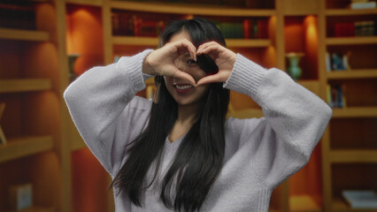 Young woman forms heart shape with hands in cozy library setting, showcasing indoor warmth and...