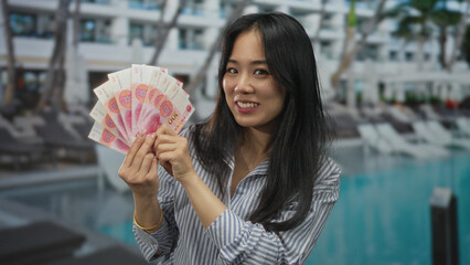 Young woman holding chinese yuan banknotes with excitement at a resort pool, showcasing financial...
