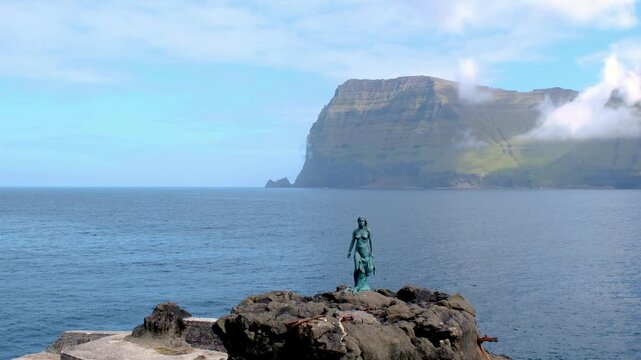 A tranquil setting on Kalsoy Island showcases dramatic cliffs and calming waters, with a striking statue overlooking the ocean. Seal woman Selkie, The Statue of Kopakonan. The Seal woman