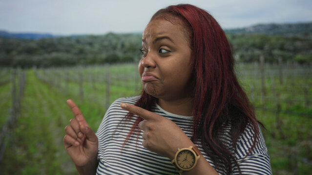 African american woman points finger at lush forest vineyard rows while looking sideways; curiosity. - Powered by Adobe