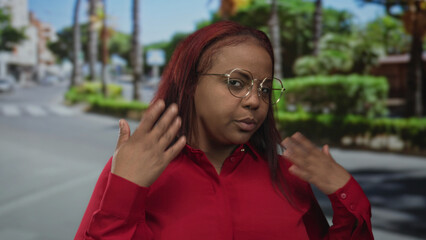 Woman in red shirt fans her face with hand on sunlit urban street lined with palm trees; heat...