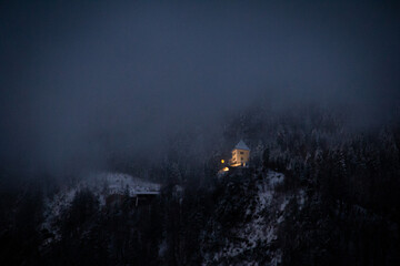 Fototapeta premium A warm light glows from a cabin tucked in snowy mountains, surrounded by mist and forest darkness. A serene and atmospheric winter escape in the wilderness.