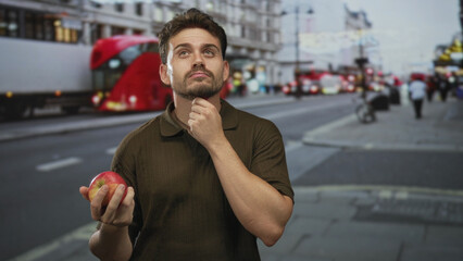 Man holding apple with hand on chin on busy city street, red bus blurred behind him and pedestrians...