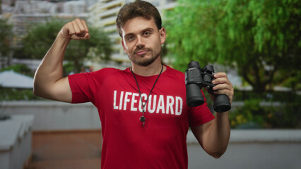 Man lifeguard wearing red shirt and whistle holds binoculars and flexes arm in front of apartment...