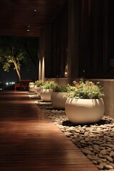 Fototapeta premium A serene outdoor walkway at night, illuminated by spotlights, featuring large white planters filled with vibrant plants, and a wooden deck.