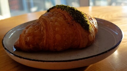 Appetizing croissant with pistachio sprinkles on a plate standing on a wooden table. In the background - a cozy cafe atmosphere with a brick wall and a window. 
