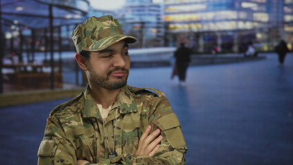 Hispanic soldier in camouflage uniform with dog tags stands with arms crossed on a city street;...