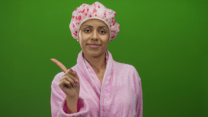 Woman in a pink bathrobe and shower cap pointing upward with one finger against an isolated green background, conveying a message of emphasis or importance.