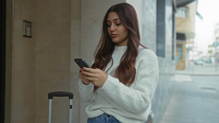 Woman using smartphone on urban street with luggage wearing sweater and jeans showcasing travel lifestyle and technology outdoors.