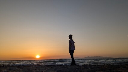 Fototapeta premium Silhouette of a girl at the beach during sunset with sea waves and mountains in the background