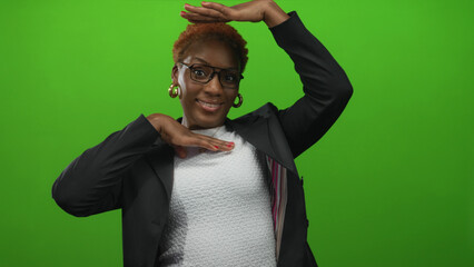 Young african american woman with glasses and hoop earrings wearing black blazer and white top arms outstretched in green studio; confidence.