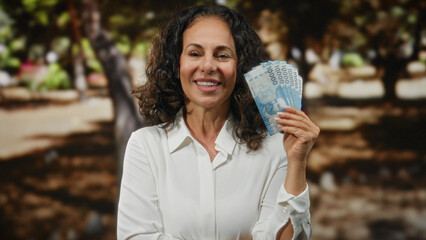 Woman smiling while holding chilean pesos in a park, showcasing currency with a joyful expression...