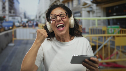 Woman celebrating with headphones and smartphone at construction site displaying excitement amidst busy urban street background.