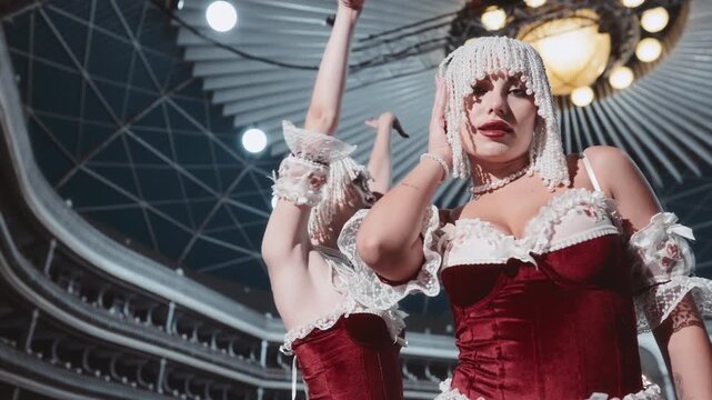 Two glamorous women in burlesque outfits pose on an empty theater stage. They are wearing red corsets with lace, pearl headdresses, and pearl necklaces.