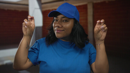 Woman shows air quotes gesture with hands in an unfinished brick building interior wearing blue cap...