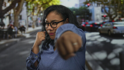 Woman wearing glasses and blue shirt with raised fists in boxing stance on city street crosswalk;...