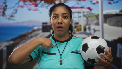 Woman referee in turquoise jersey holds soccer ball on seaside promenade while pointing finger to chest; confidence.