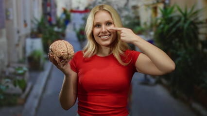 Woman smiling outdoors holding a brain model while touching her nose on a vibrant city street.