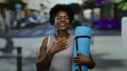 Woman holding yoga mat smiling confidently in urban street setting, embodying fitness and wellness...