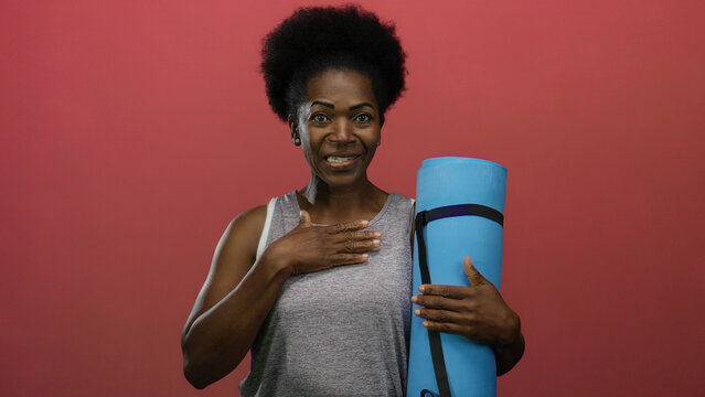 Woman holding yoga mat against red background, looking cheerful and confident.