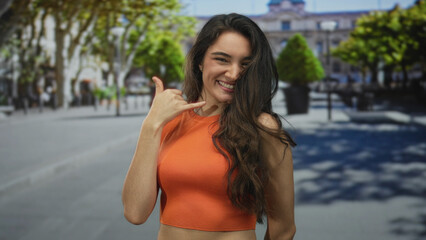 Woman makes call me gesture with hand to ear and pinky to lips in city street scene; cheerful...