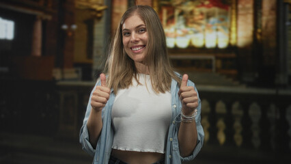 Young blonde woman wearing casual denim shirt and white top giving thumbs up gesture in an ornate catholic church interior; approval.
